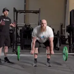 Man med skägg förbereder sig för en romanian deadlift med en skivstång. En tränare står bredvid och instruerar.