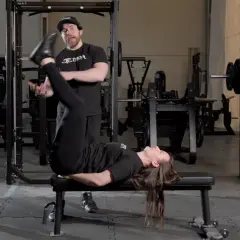 En kvinna utför raka benlyft på en bänk i ett gym, med en tränare som observerar.