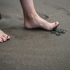 Nära en sten på stranden, en bar fot med rött nagellack och sand mellan tårna. En annan fot med eversion fot syns i bakgrunden.