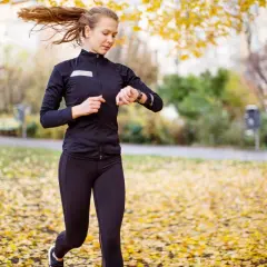 En kvinna springer i en park täckt av höstlöv och kollar sin klocka, kanske för att se hur lång tid det tar att springa 5 km.