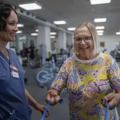 En sjuksköterska assisterar en äldre kvinna med medicinsk rehabilitering i ett gym. Kvinnan ler glatt medan hon tränar med ett blått gummiband.