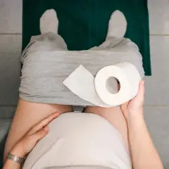 Gravid kvinna med hård i magen sitter på toaletten med en rulle toalettpapper.