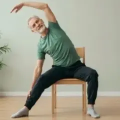 Äldre man med grått hår och skägg sitter på en stol och sträcker sig åt sidan. Han verkar ha dålig balans när han gör övningen.