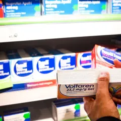 En hand håller upp en förpackning Voltaren gel med diklofenak nsaid. I bakgrunden syns hyllor med fler läkemedel.