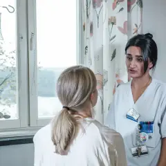 Sjuksköterska pratar med patient om biverkning kortison. Droppställning och medicinsk utrustning syns i bakgrunden.