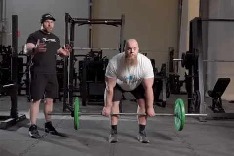 Man med skägg förbereder sig för en romanian deadlift med en skivstång. En tränare står bredvid och instruerar.