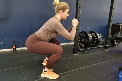 En kvinna med håret uppsatt i en knut utför squats svenska i ett gym. Hon bär bruna träningskläder och orangea skor. I bakgrunden syns en skivstångsställning med vikter.