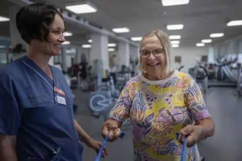 En sjuksköterska assisterar en äldre kvinna med medicinsk rehabilitering i ett gym. Kvinnan ler glatt medan hon tränar med ett blått gummiband.