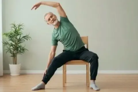 Äldre man med grått hår och skägg sitter på en stol och sträcker sig åt sidan. Han verkar ha dålig balans när han gör övningen.