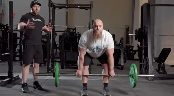Man med skägg förbereder sig för en romanian deadlift med en skivstång. En tränare står bredvid och instruerar.