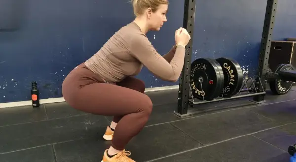 En kvinna med håret uppsatt i en knut utför squats svenska i ett gym. Hon bär bruna träningskläder och orangea skor. I bakgrunden syns en skivstångsställning med vikter.