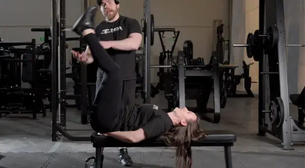 En kvinna utför raka benlyft på en bänk i ett gym, med en tränare som observerar.