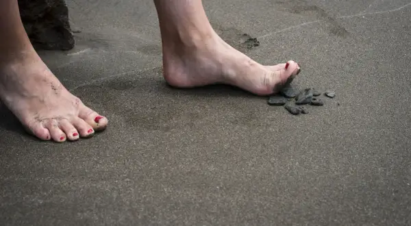 Nära en sten på stranden, en bar fot med rött nagellack och sand mellan tårna. En annan fot med eversion fot syns i bakgrunden.