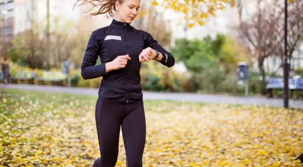 En kvinna springer i en park täckt av höstlöv och kollar sin klocka, kanske för att se hur lång tid det tar att springa 5 km.