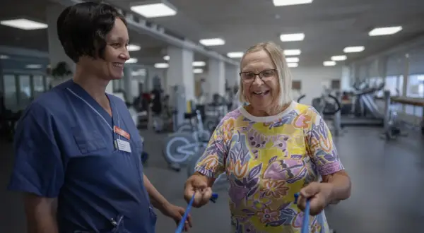 En sjuksköterska assisterar en äldre kvinna med medicinsk rehabilitering i ett gym. Kvinnan ler glatt medan hon tränar med ett blått gummiband.