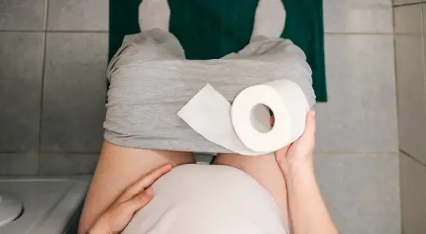 Gravid kvinna med hård i magen sitter på toaletten med en rulle toalettpapper.