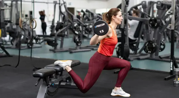 Kvinna utför bulgariska utfallssteg med skivstång på axlarna i ett gym.