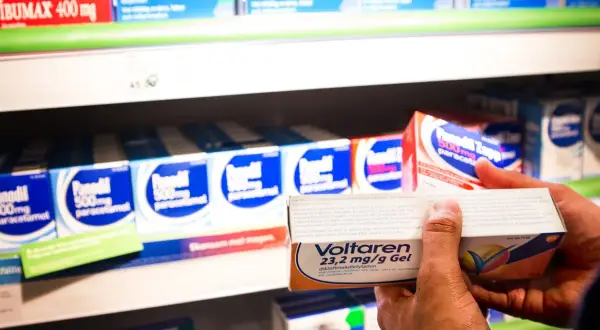 En hand håller upp en förpackning Voltaren gel med diklofenak nsaid. I bakgrunden syns hyllor med fler läkemedel.
