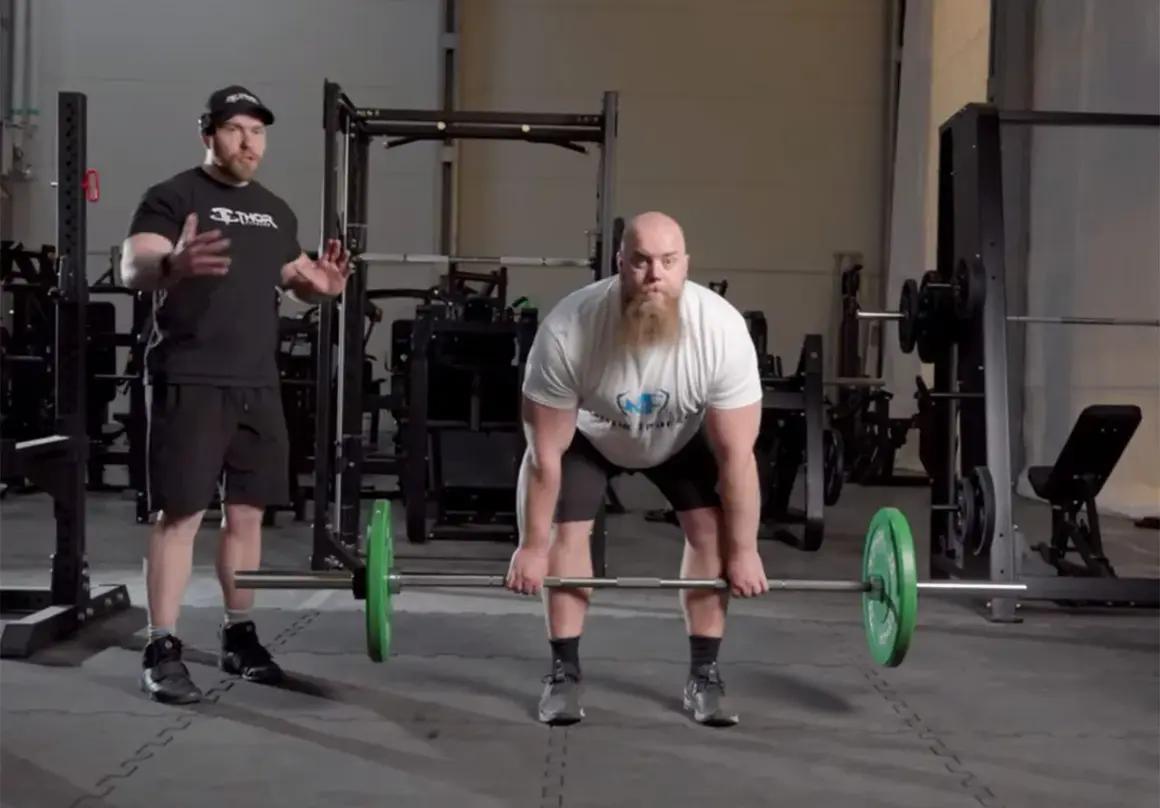 Man med skägg förbereder sig för en romanian deadlift med en skivstång. En tränare står bredvid och instruerar.