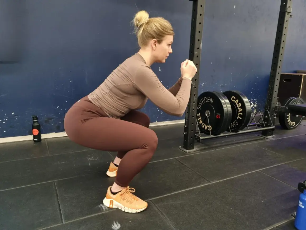 En kvinna med håret uppsatt i en knut utför squats svenska i ett gym. Hon bär bruna träningskläder och orangea skor. I bakgrunden syns en skivstångsställning med vikter.
