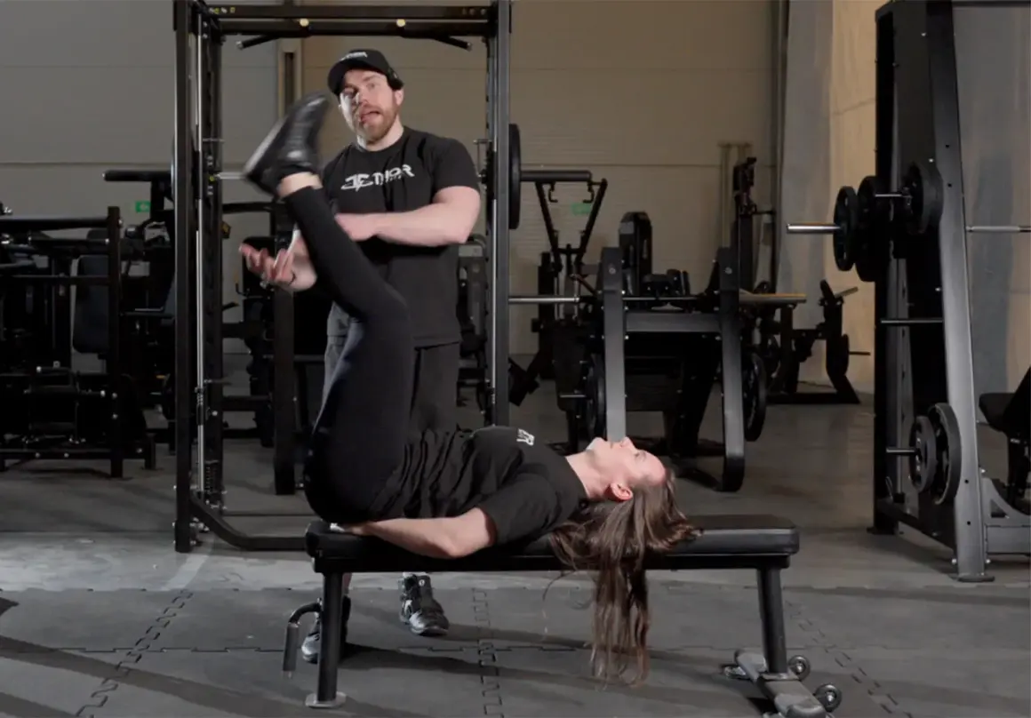En kvinna utför raka benlyft på en bänk i ett gym, med en tränare som observerar.