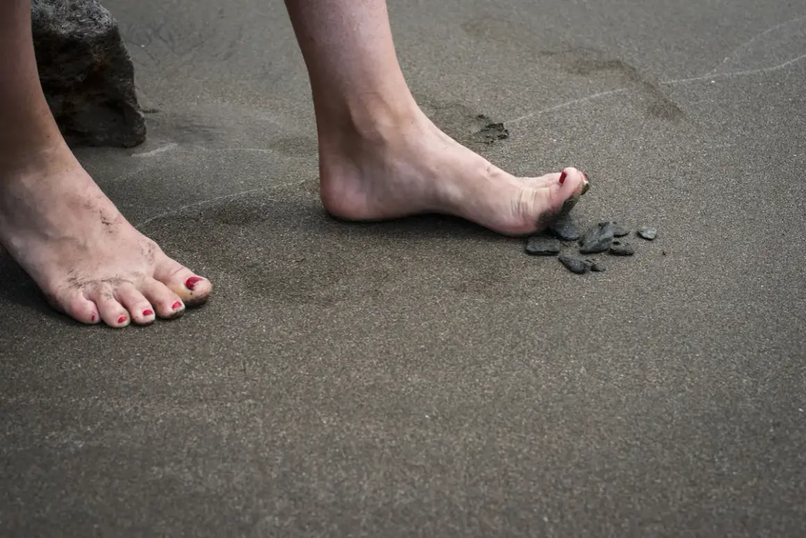 Nära en sten på stranden, en bar fot med rött nagellack och sand mellan tårna. En annan fot med eversion fot syns i bakgrunden.