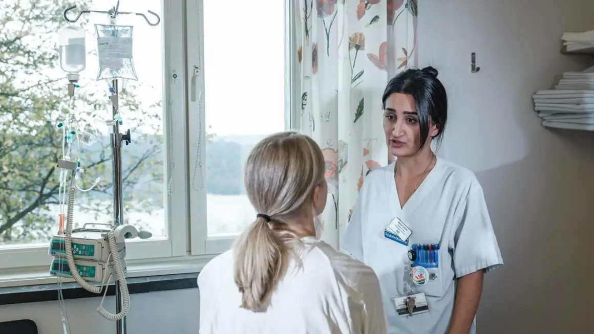 Sjuksköterska pratar med patient om biverkning kortison. Droppställning och medicinsk utrustning syns i bakgrunden.
