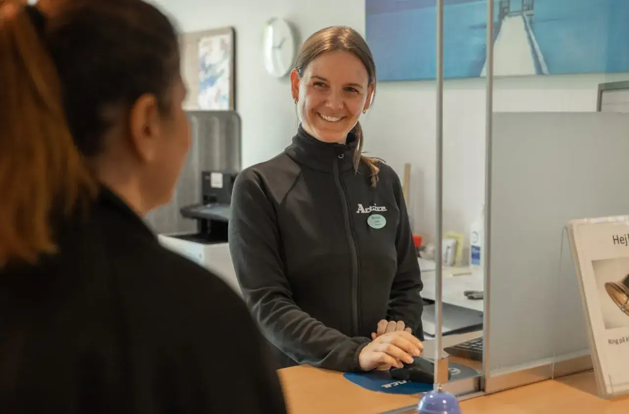 En leende kvinna bakom en reception förklarar skillnad på arbetsterapeut och fysioterapeut.