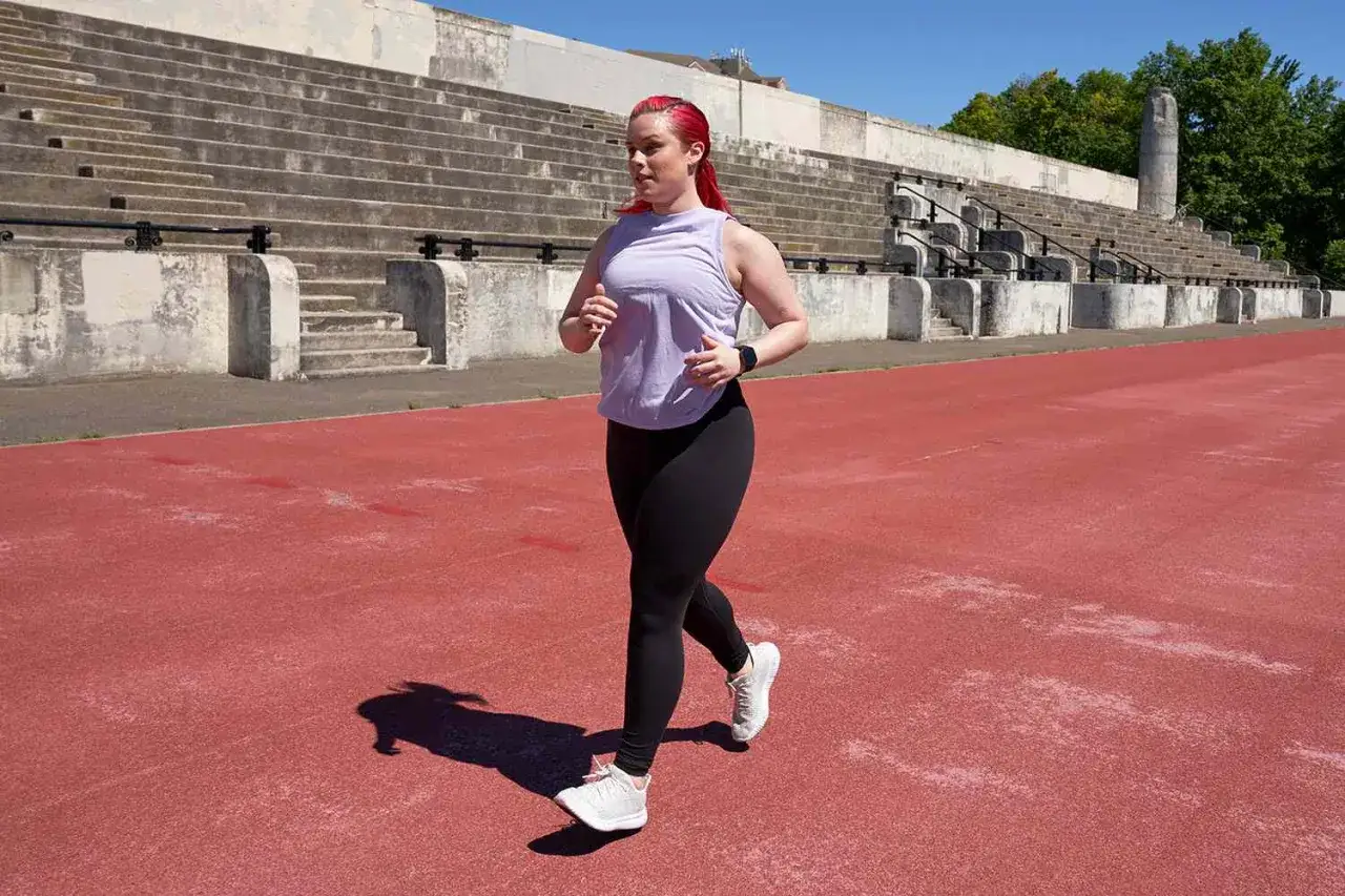 Kvinna med rött hår springer på en röd löparbana vid en gammal arena, redo för sitt cooper test.