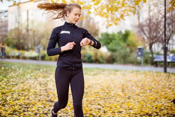 En kvinna springer i en park täckt av höstlöv och kollar sin klocka, kanske för att se hur lång tid det tar att springa 5 km.
