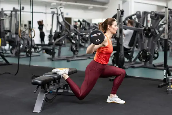 Kvinna utför bulgariska utfallssteg med skivstång på axlarna i ett gym.