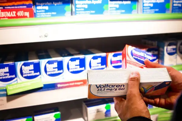 En hand håller upp en förpackning Voltaren gel med diklofenak nsaid. I bakgrunden syns hyllor med fler läkemedel.